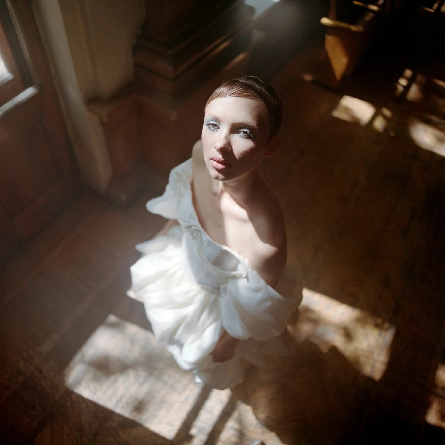 A woman with short hair sitting on a wooden chair, looking up at the camera with sunlight casting shadows on her face and dress.