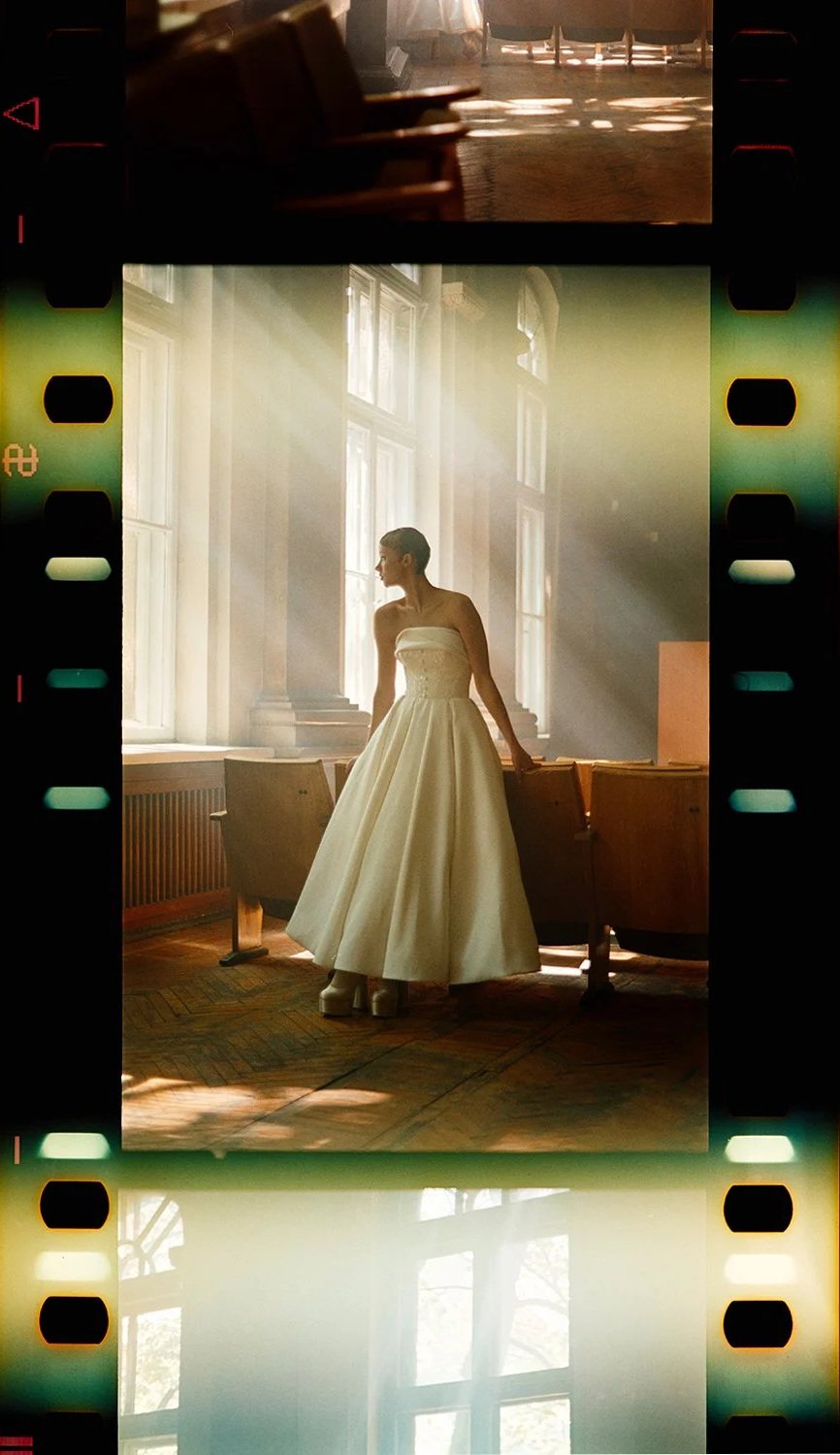 A woman in a strapless white gown standing in a sunlit room with large windows, wooden floors, and vintage chairs, creating a soft, elegant atmosphere.