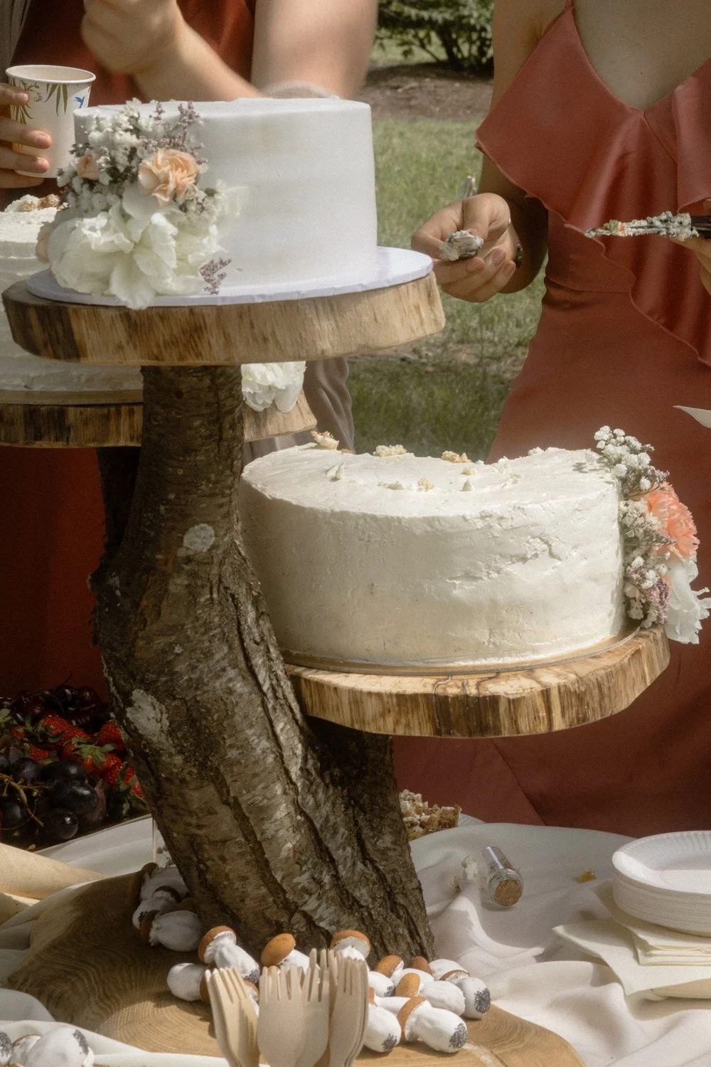 Two white frosted wedding cakes with floral decorations on a rustic wooden cake stand at an outdoor event. One cake is on a higher tier, and the other is on a lower tier. There are also some small desserts and cups on the table.
