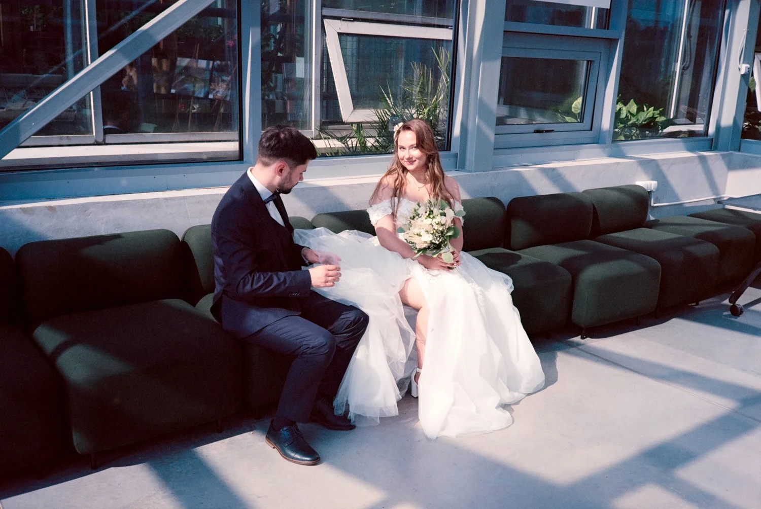 A bride and groom sitting on a green couch inside a modern building with large windows, with the bride holding a bouquet of white flowers and the groom adjusting his sleeves.