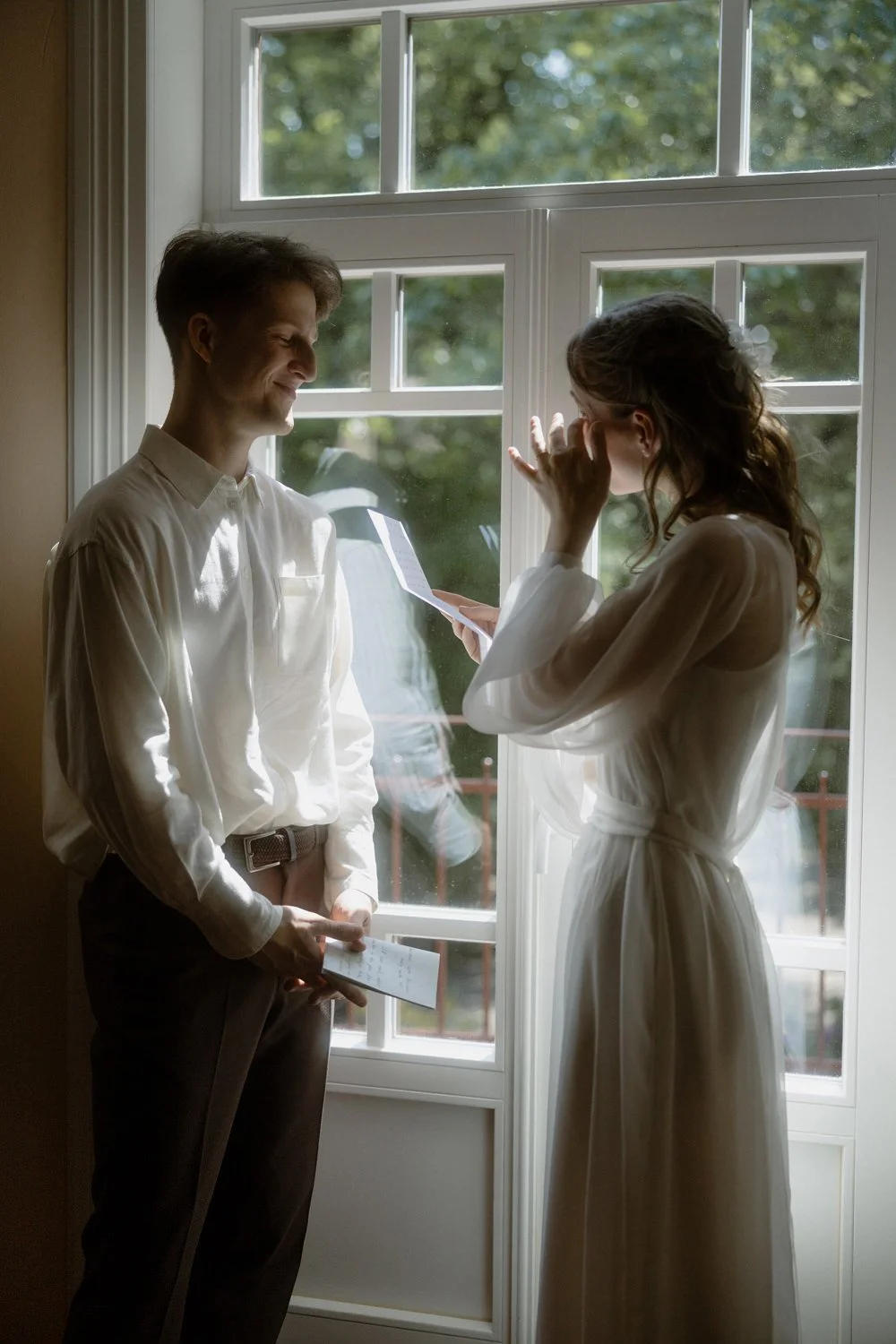 A young man and woman stand indoors near a large window with greenery outside. The woman appears emotional, holding her hand to her face, while the man smiles gently. She is wearing a white dress with a floral hair accessory, and he is dressed in a w