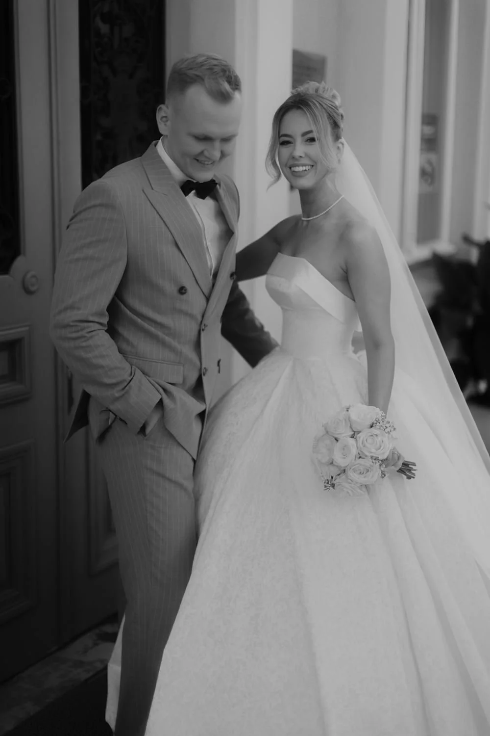 A bride and groom on their wedding day, smiling and posing indoors, the bride holding a bouquet of roses.