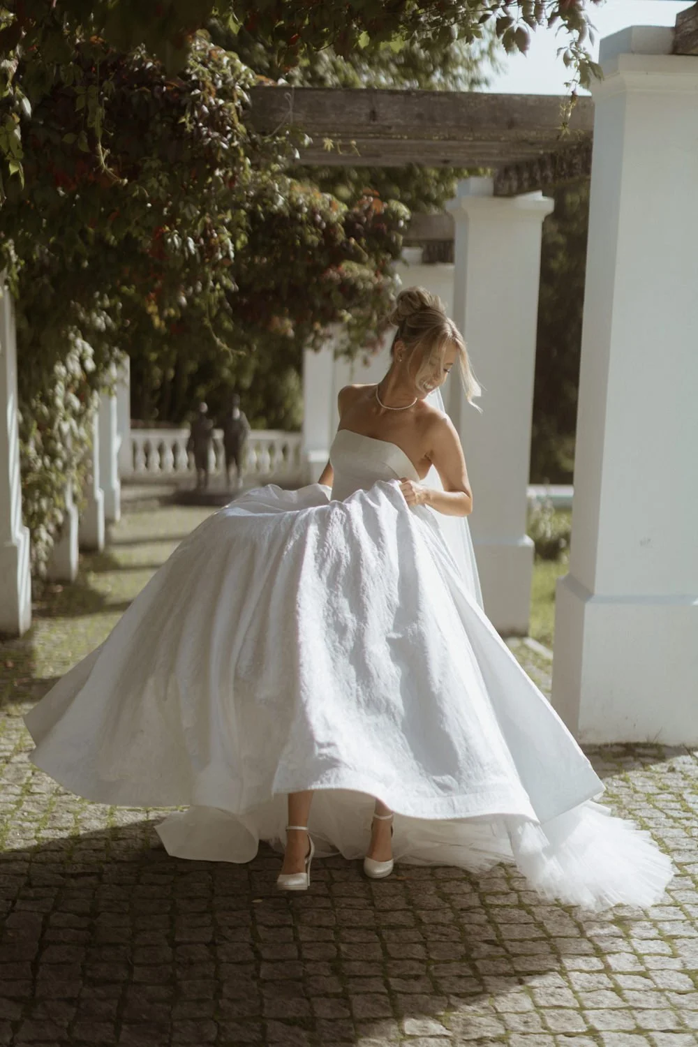 A bride in a white wedding gown standing outdoors on a cobblestone path. She is holding her dress and looking down. The background shows white columns, greenery, and two people walking in the distance.