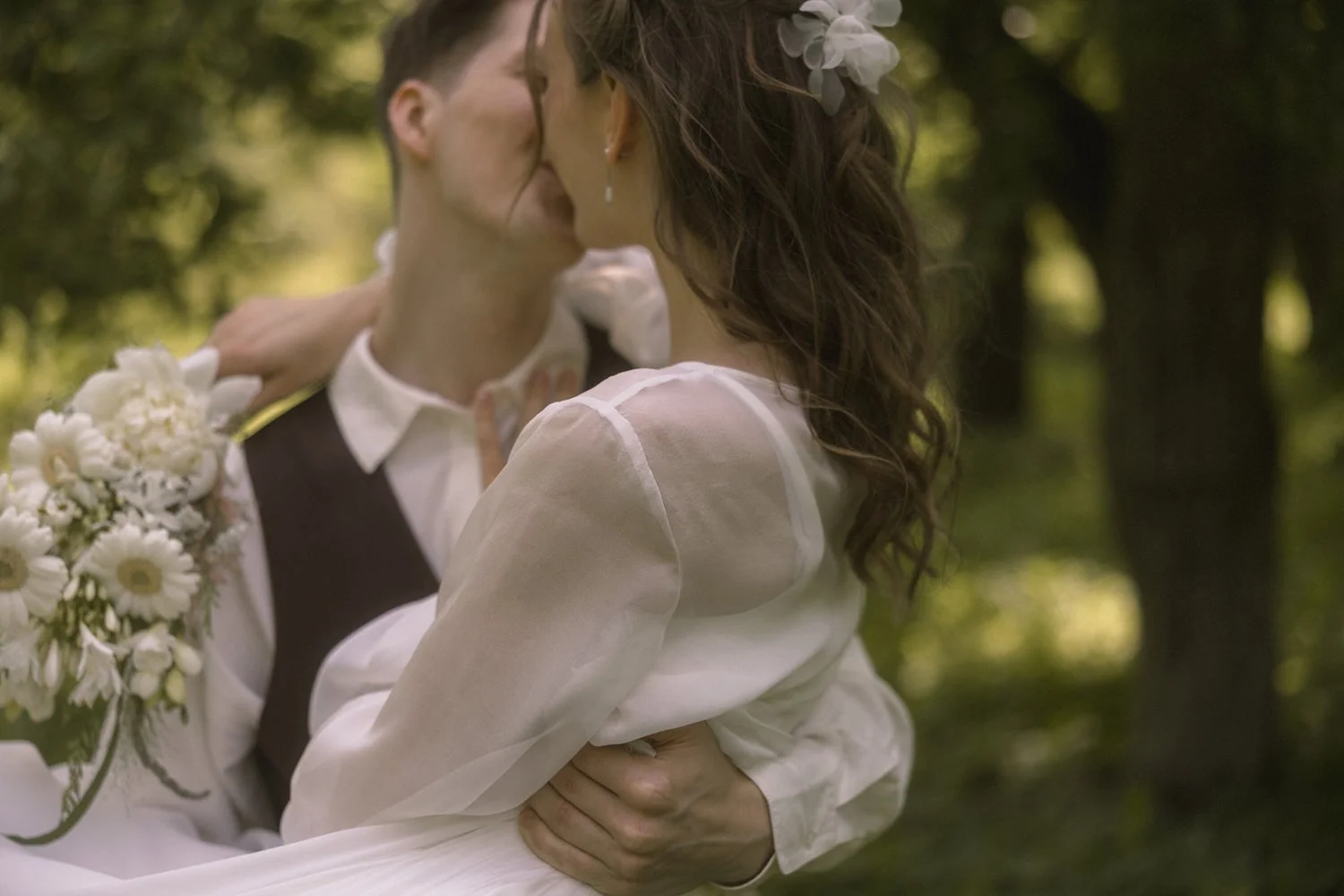 A woman and a man sharing a kiss outdoors in a forest setting, with the woman holding a bouquet of white flowers.