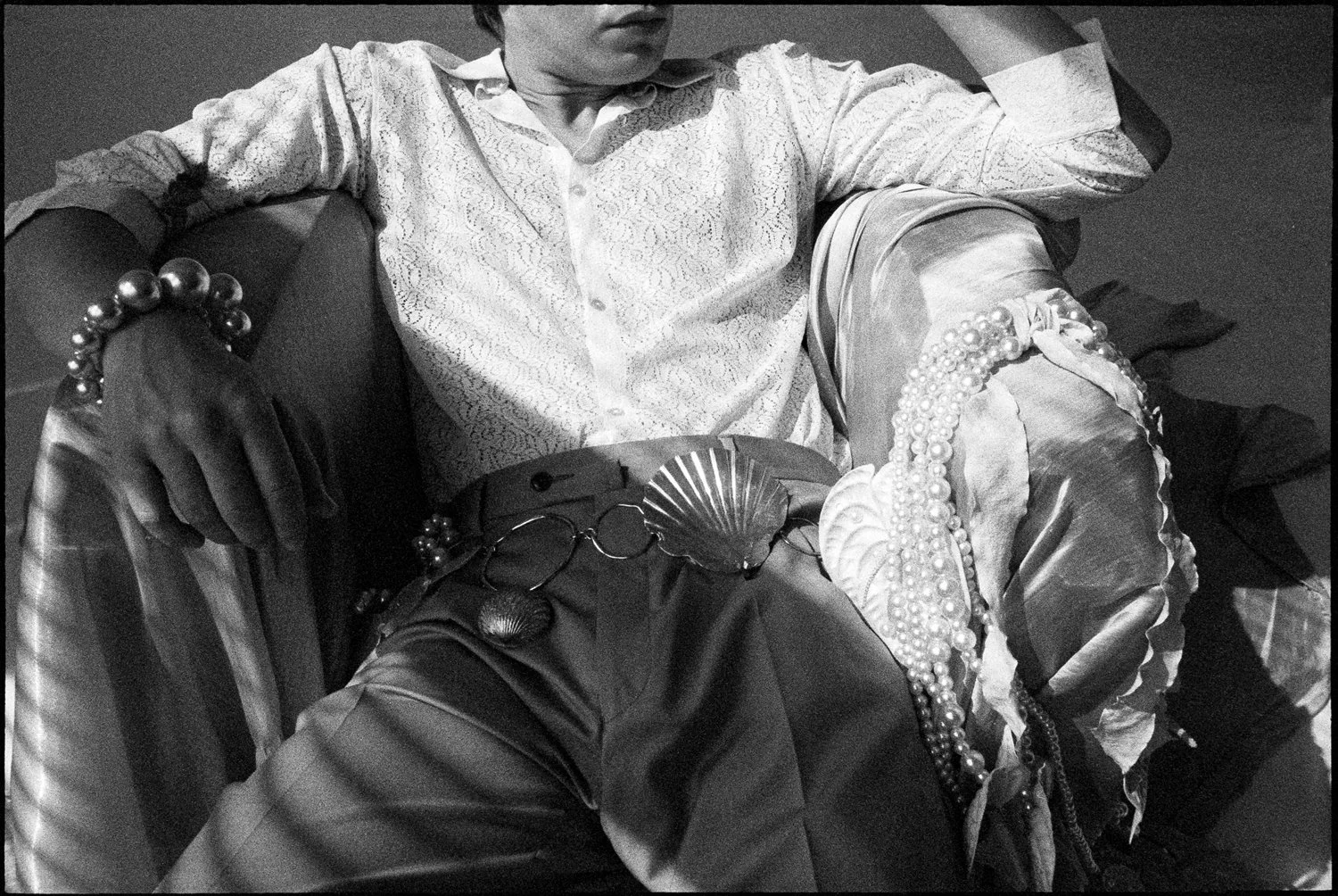 Black and white photo of a person sitting with crossed legs, adorned with multiple pearl necklaces, a pearl bracelet, and earrings. The person is wearing a button-up shirt with embroidery or lace details and leather pants, with glasses hanging from a