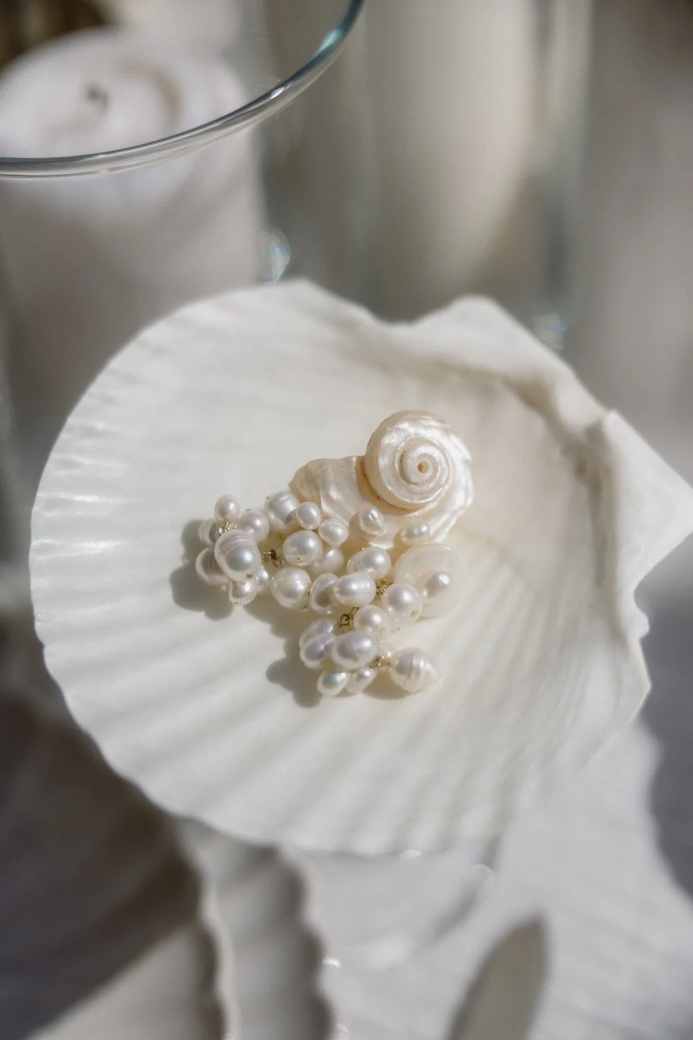 Pearl jewelry arranged on a white seashell with a glass container in the background.