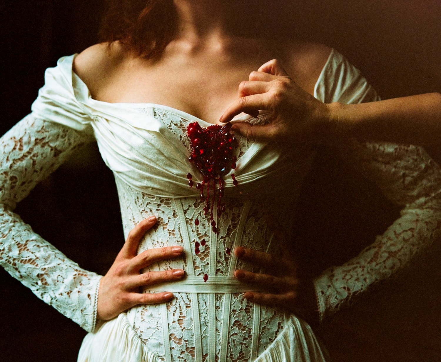 Close-up of a woman wearing an elegant white lace dress with a large red embroidered heart on the chest, surrounded by beads, and with her hands on her hips.