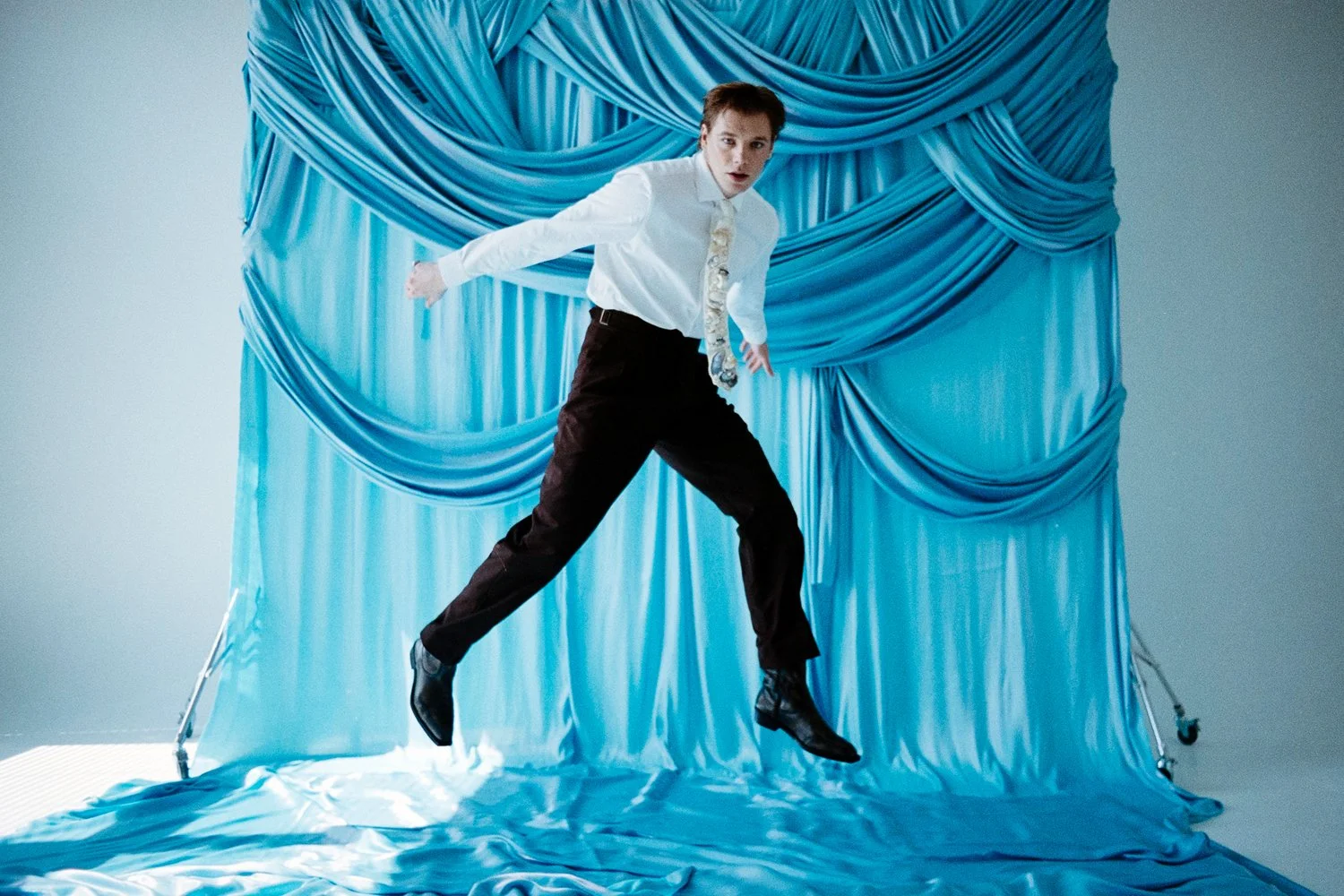 A man wearing a white shirt and black pants appears to be floating or falling in the air in front of a blue satin curtain backdrop.