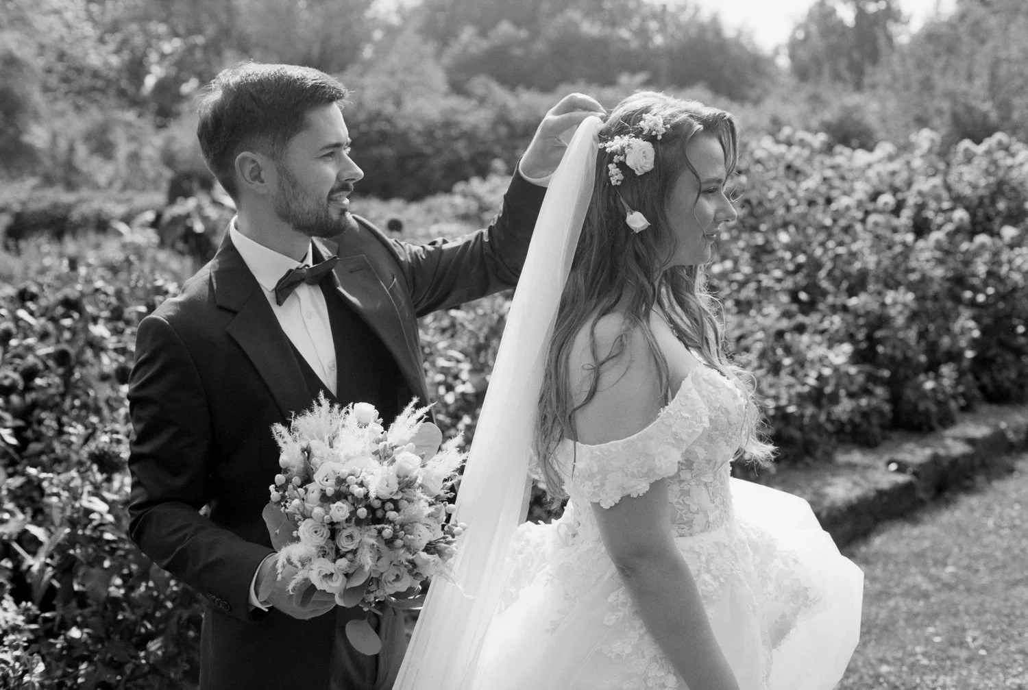 A black and white photo of a groom adjusting a bride's veil during an outdoor wedding. The groom is holding a bouquet, and the bride is wearing an off-shoulder lace wedding gown with flowers in her hair, standing in a garden.