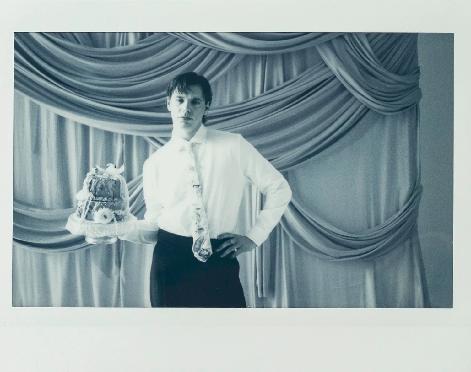 A young man in a white shirt and tie holding a decorated cake on a cake stand, standing in front of draped curtains.