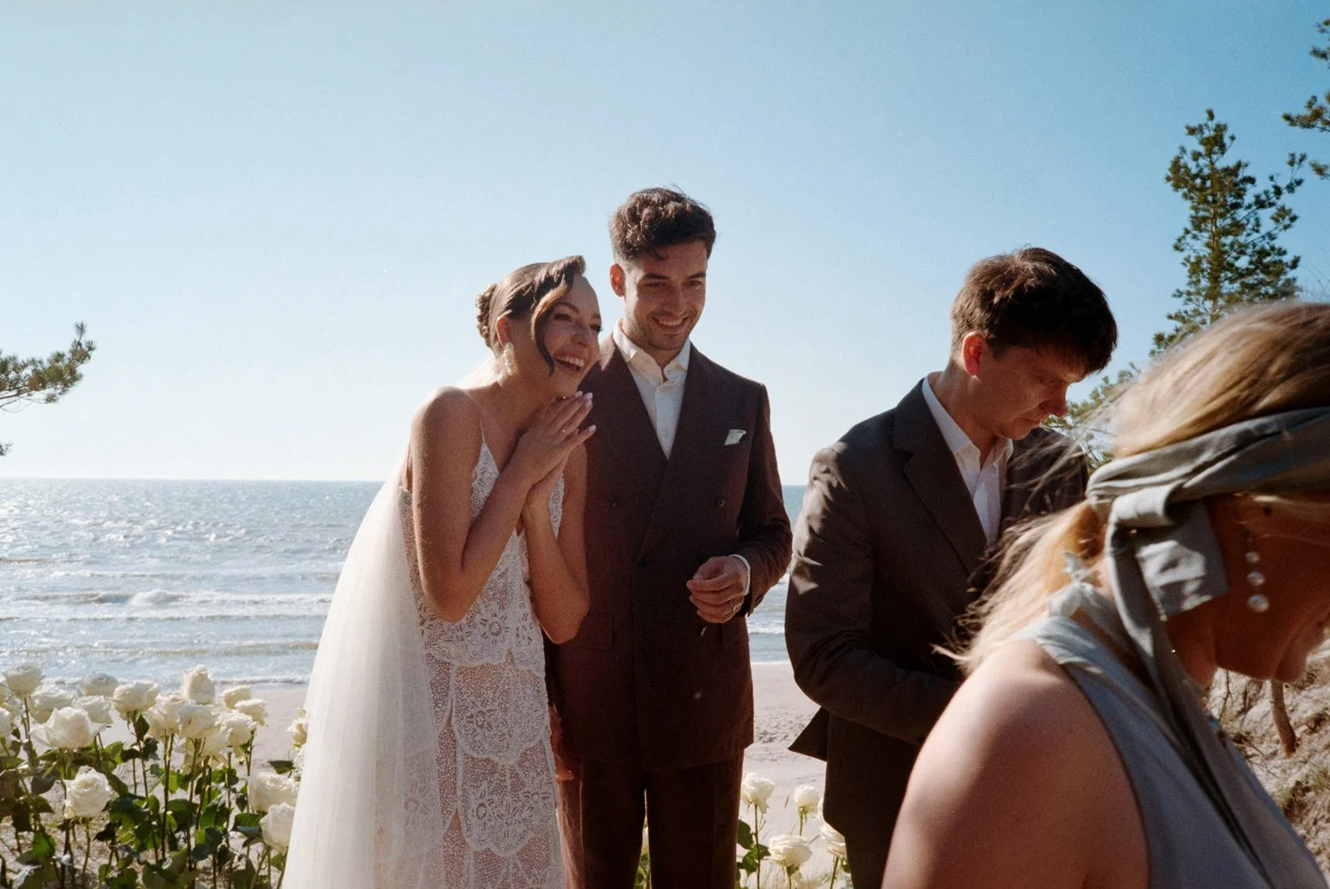 A group of people at a beach wedding, including a bride in a lace wedding dress smiling and covering her mouth, two men in suits, and a woman with blonde hair and a scarf, with the ocean, trees, and sky in the background.