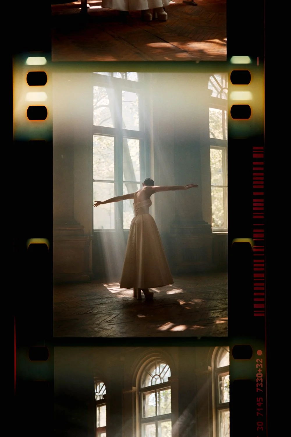 A dancer in a long, elegant dress performs in a sunlit room with large windows and wooden floors, light beams creating a dramatic effect.