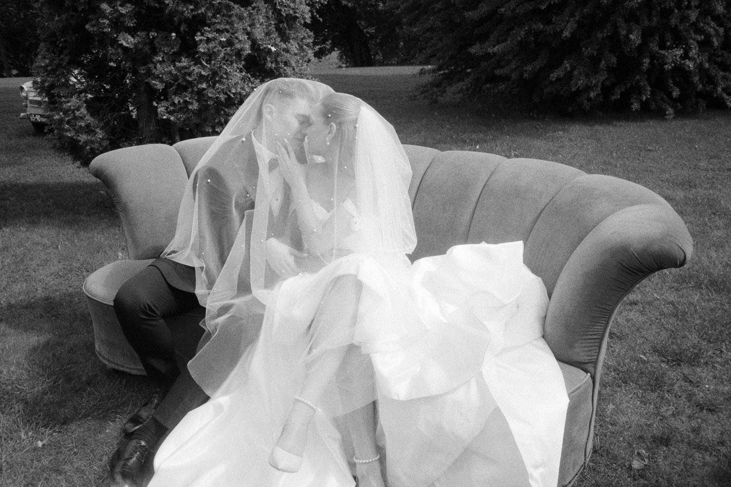A bride and groom sitting closely on a vintage sofa outdoors, sharing a kiss through a veil on a wedding day.