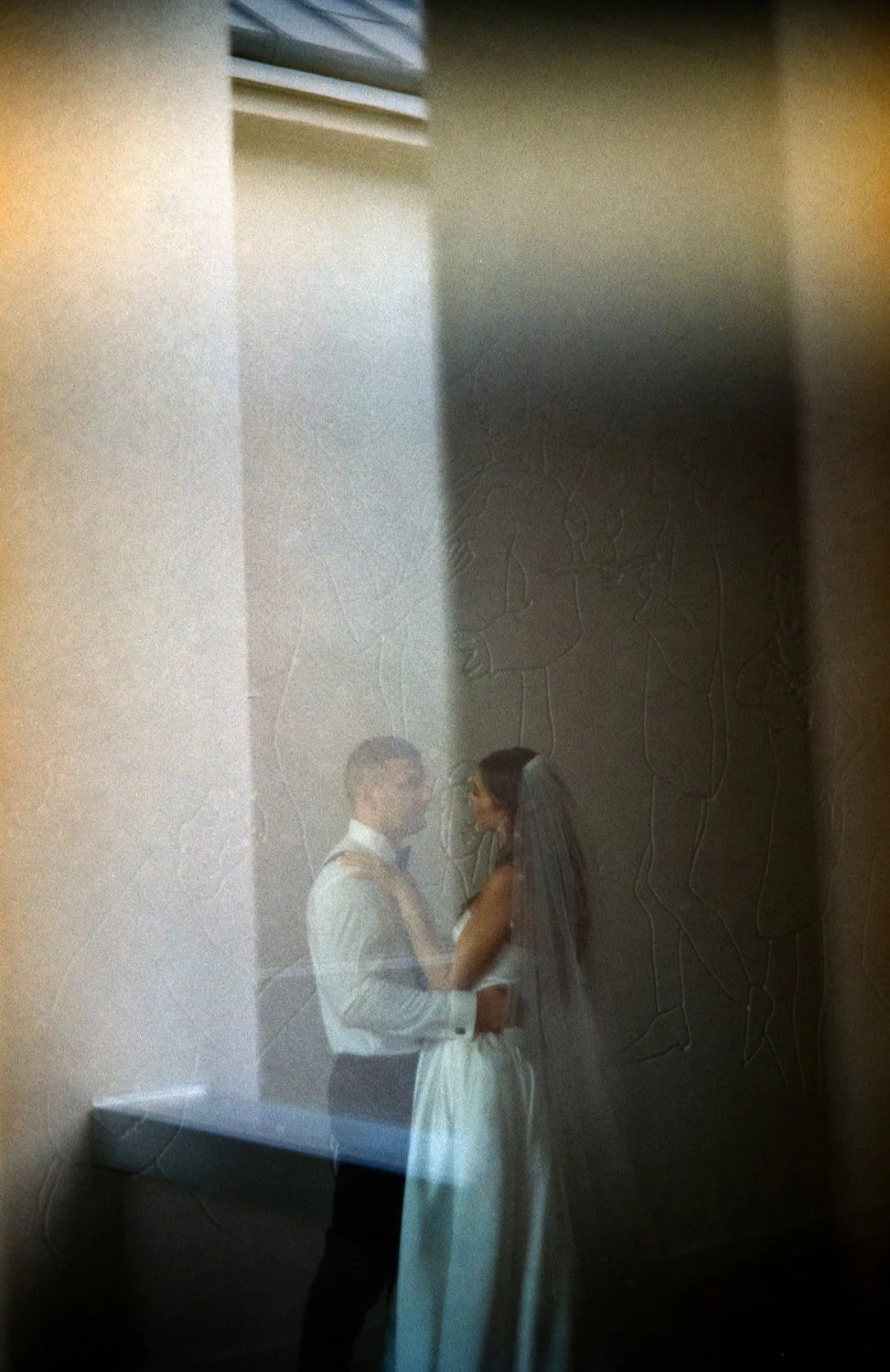 Reflection of a bride and groom standing close together on a wall with carved figures, seen through a glass surface.