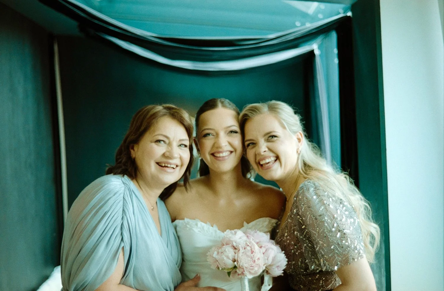 Three women smiling and celebrating, with one in a white dress holding a bouquet, likely at a wedding, in a room with teal walls.