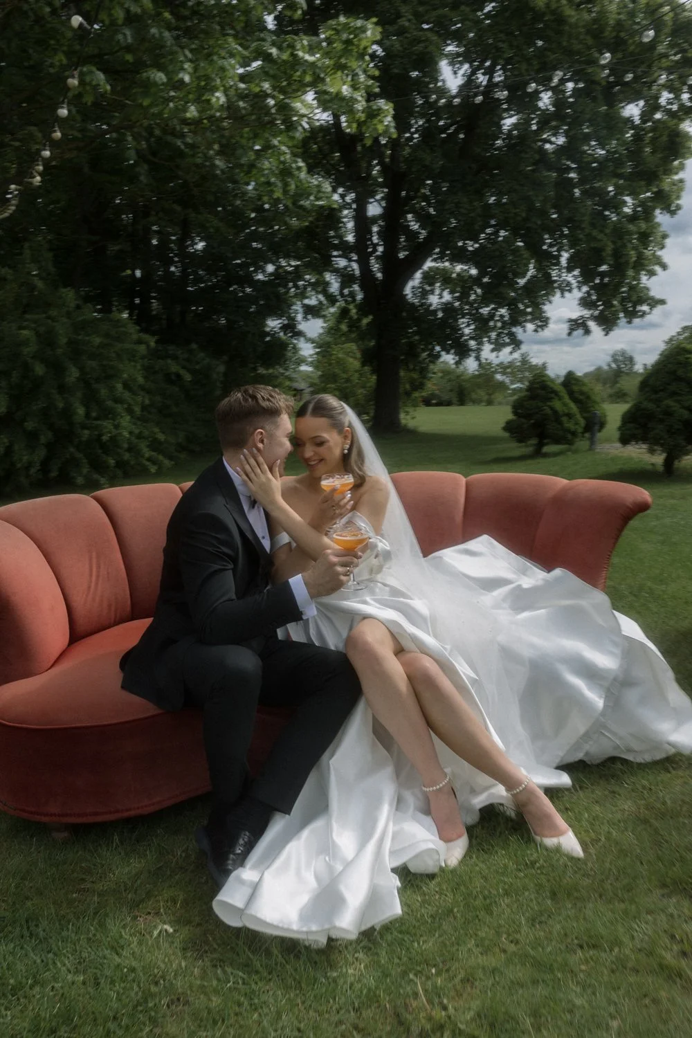 A bride and groom sitting on a vintage pink sofa outdoors, smiling and holding cocktails, in a lush green park setting, sharing an intimate moment.