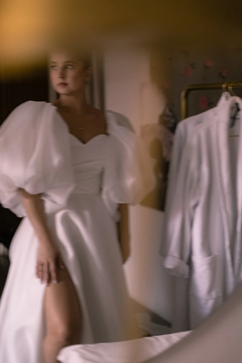 A woman in a white dress standing by a clothing rack with white garments, in a softly lit room.