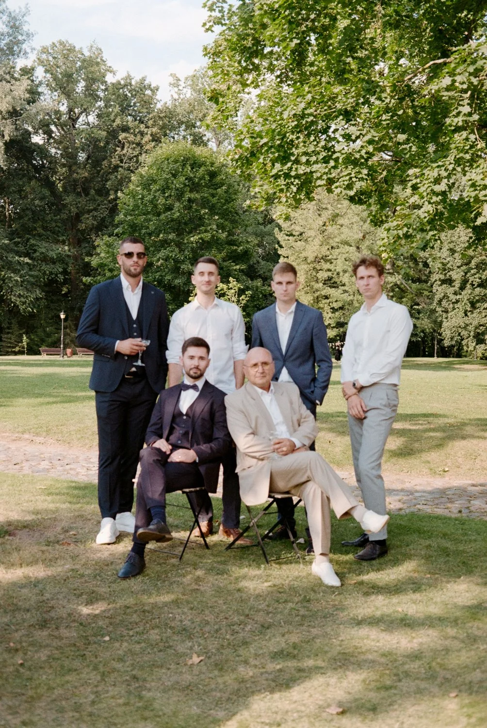 Six men dressed in formal and semi-formal attire posing outdoors in a park with trees in the background. Some are standing and two are seated in chairs, with one holding a glass.