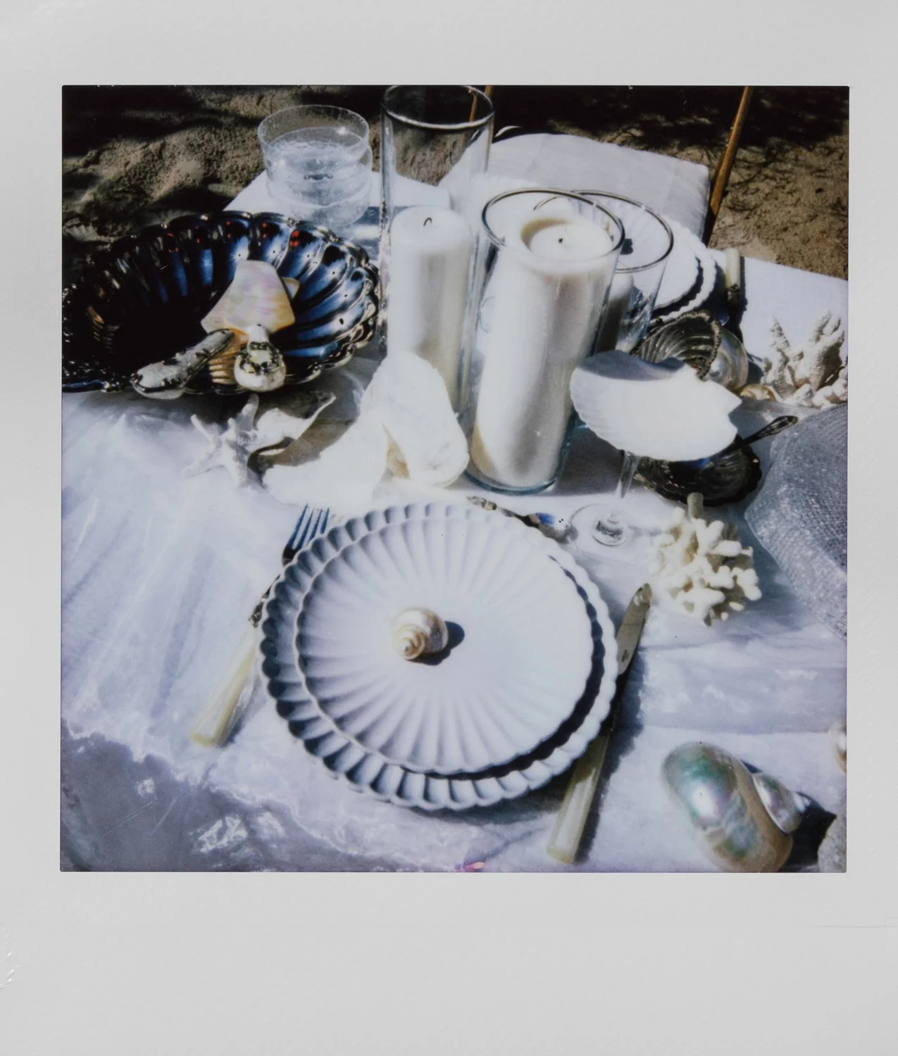 Table set with seashells, candles, glassware, and a white and blue tablecloth, in an outdoor setting.