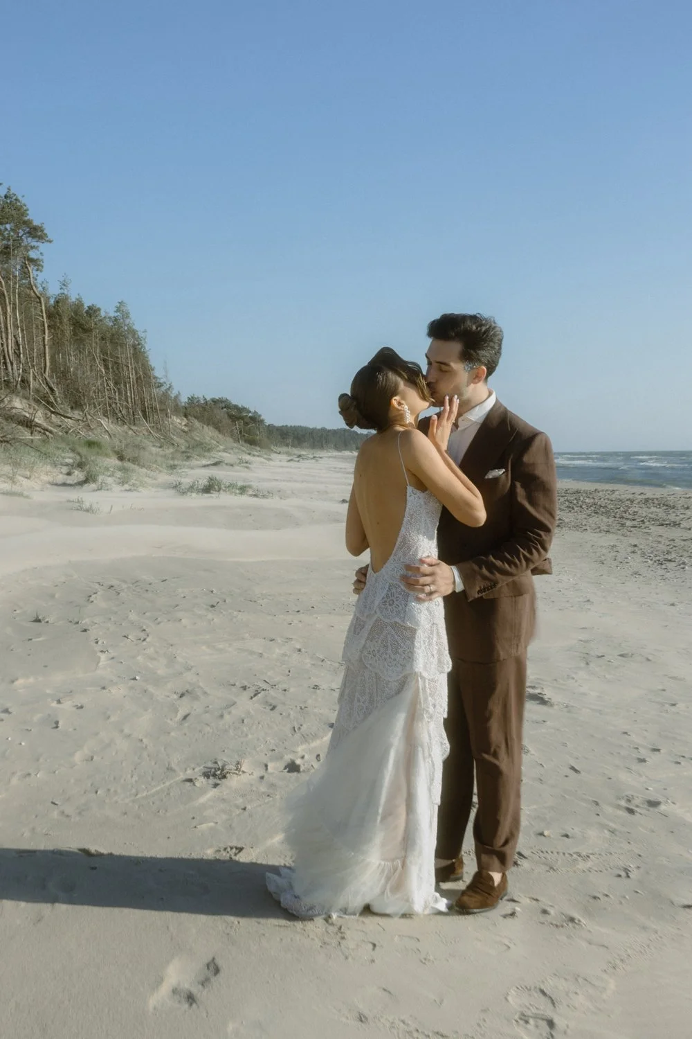 A couple dressed in wedding attire sharing a kiss on a beach during the daytime with sand dunes and blue sky in the background.