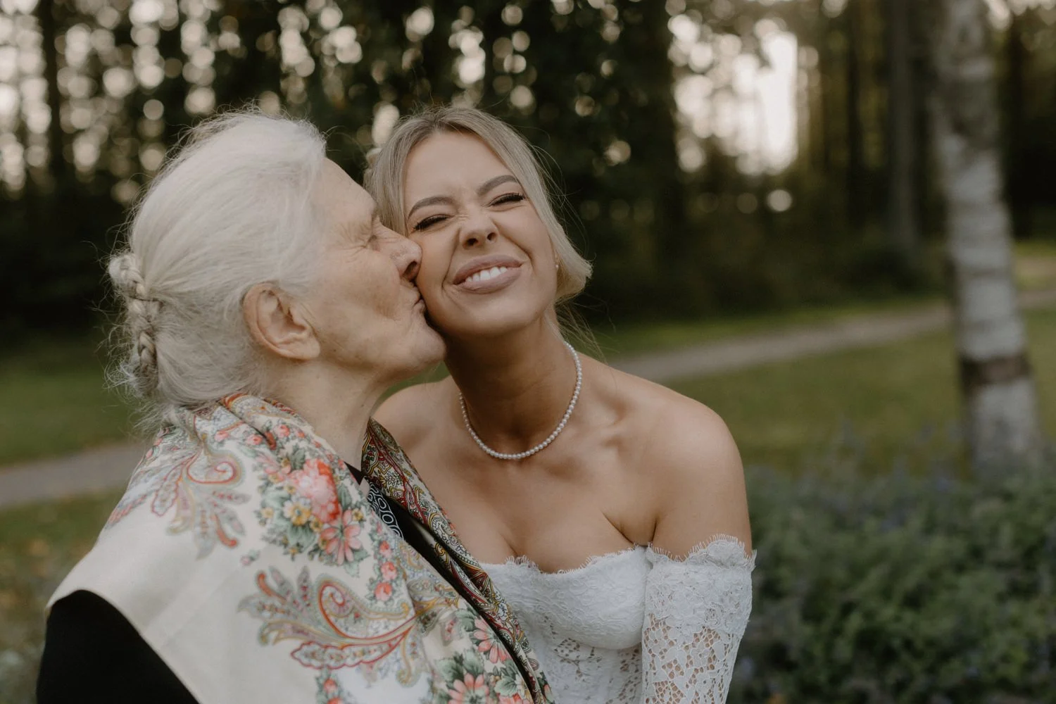 An elderly woman with white hair styled in a braid gives a kiss on the cheek to a smiling woman with blonde hair in an outdoor park setting.