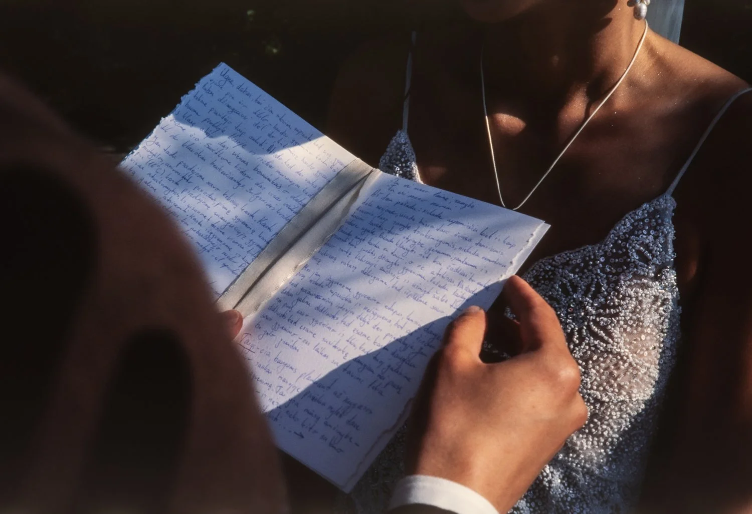 A person wearing a beaded dress is holding and reading handwritten notes in a notebook. The scene is illuminated by warm sunlight, and the person is wearing earphones.