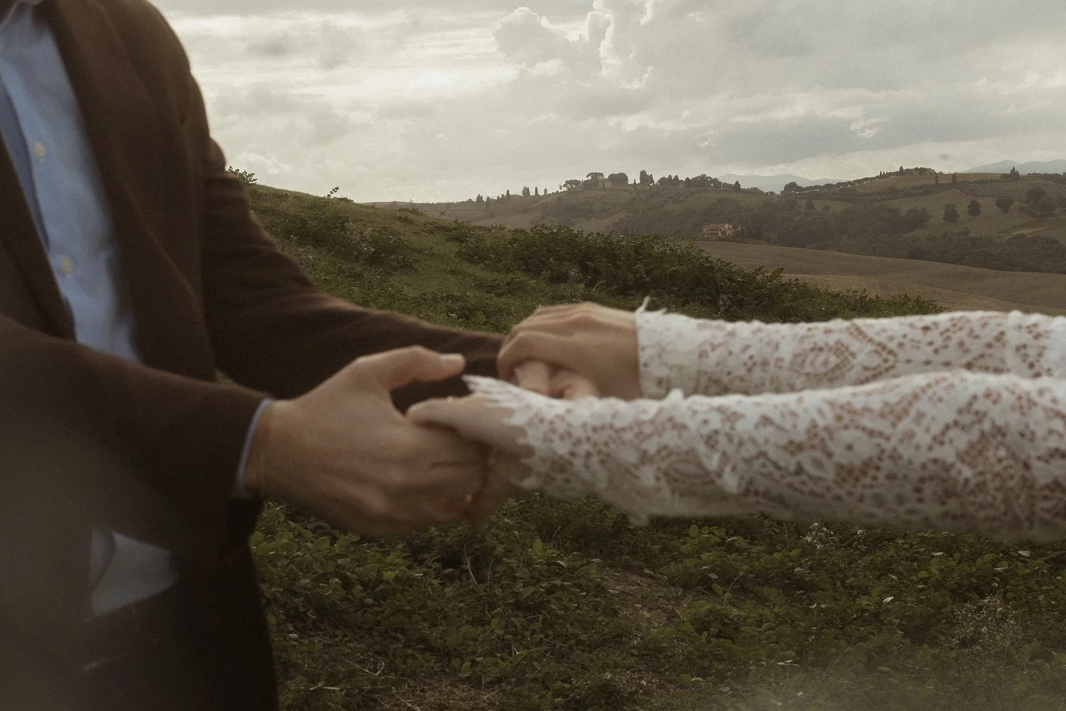 Two people holding hands in tuscany, setting with rolling hills and cloudy sky in the background.