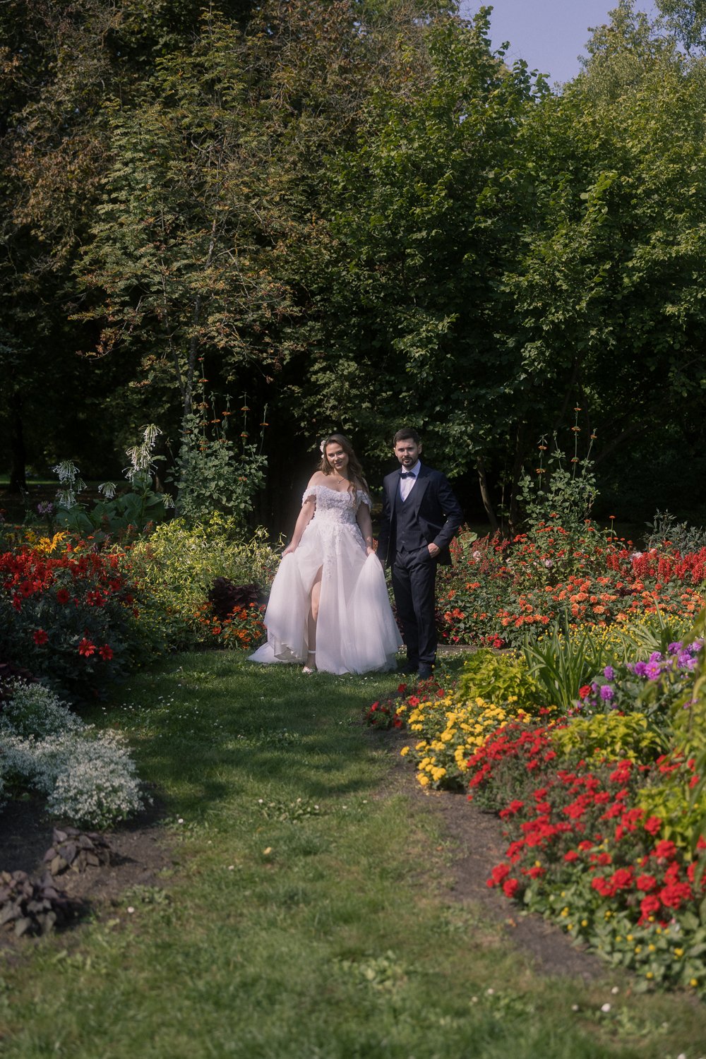 A bride and groom posing together in a garden with colorful flowers and green trees in the background.