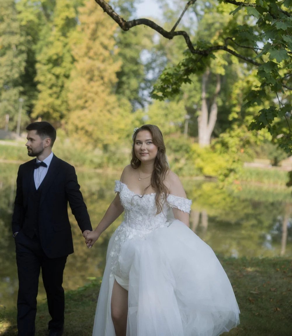 A bride in a white wedding dress holding hands with a groom in a black suit and bow tie walking outdoors near a lake, surrounded by trees with green foliage.