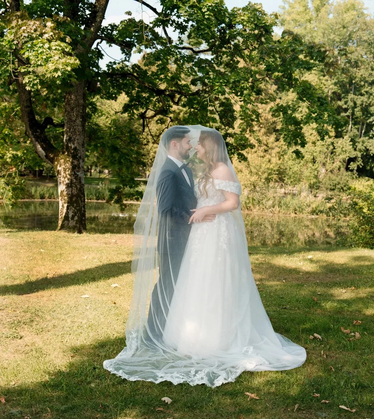 A bride and groom standing close together outdoors on grass with trees and a pond in the background, sharing a moment under a veil.