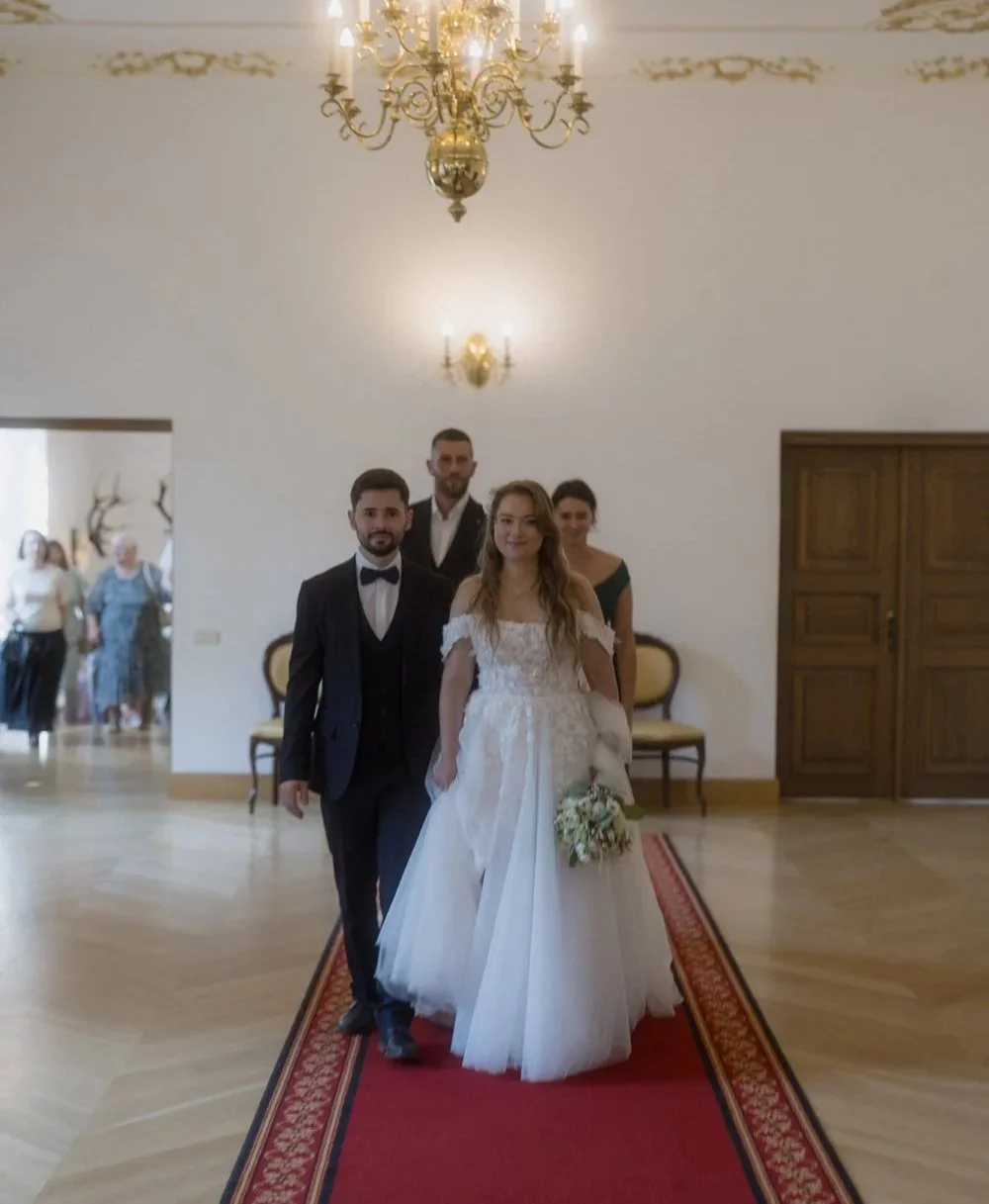 Bride and groom walking down aisle in wedding ceremony with bridesmaids and groomsmen in background, inside a decorated hall with chandelier.