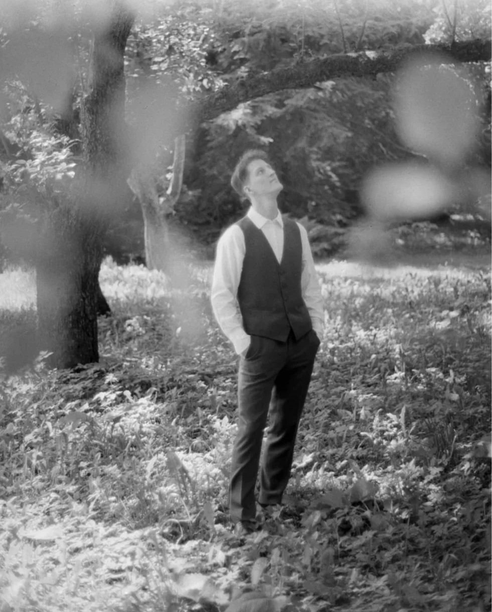 A man in a vest and slacks standing in a wooded outdoor area, looking up.