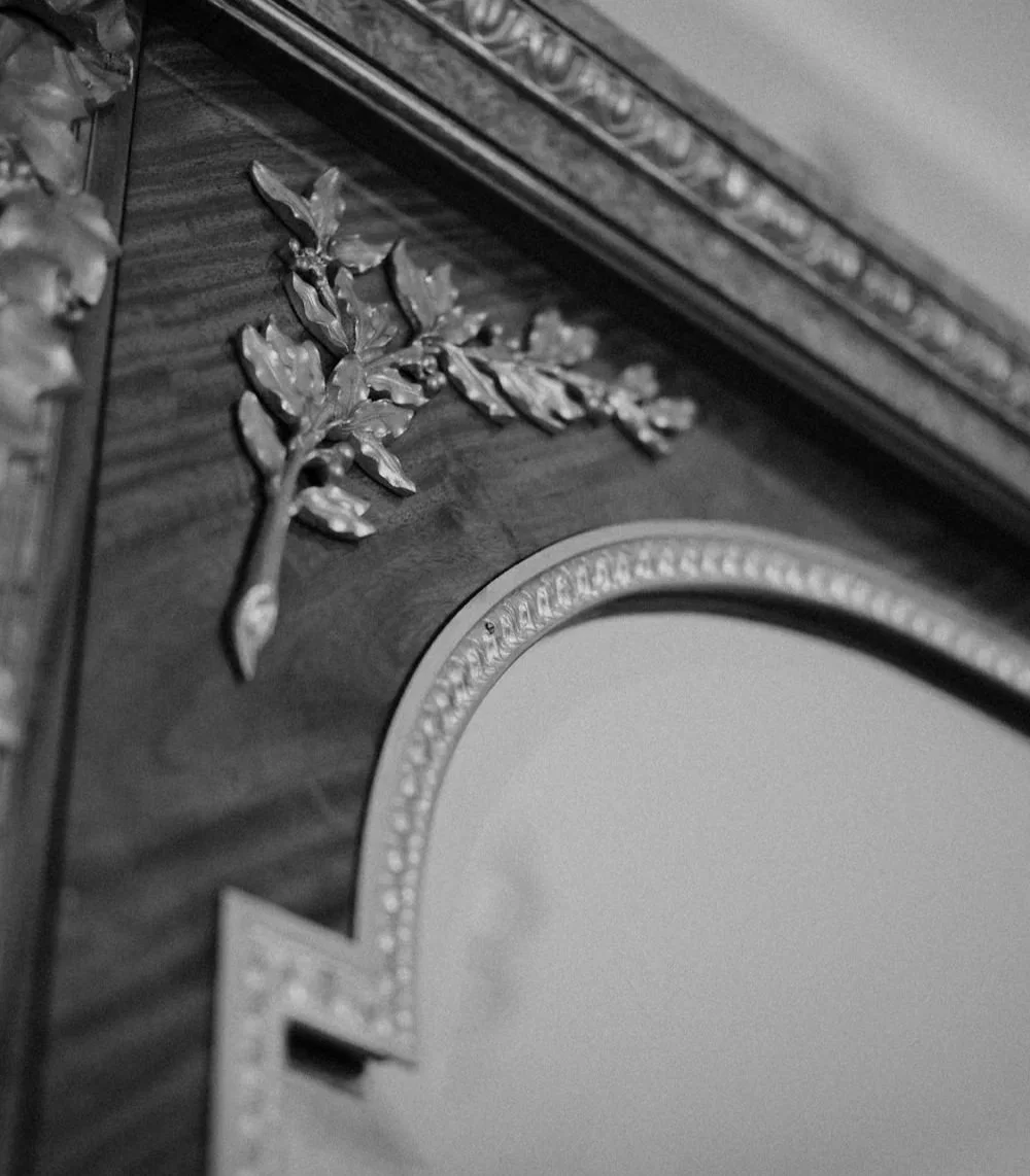 Close-up of an ornate wooden piece with a decorative metal leaf detail, part of a mirror frame.