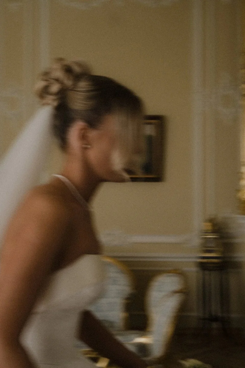 A woman with a bun hairstyle wearing a white strapless dress, possibly a wedding gown, standing in a room with beige walls and a dark framed picture.