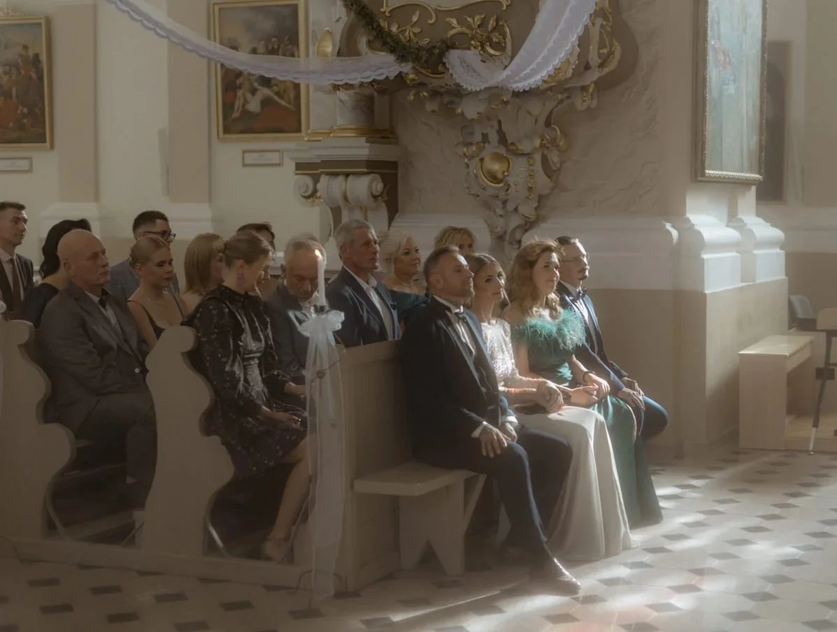 People sitting in church pews during a ceremony, possibly a wedding, inside a decorated church with ornate wall details and paintings.
