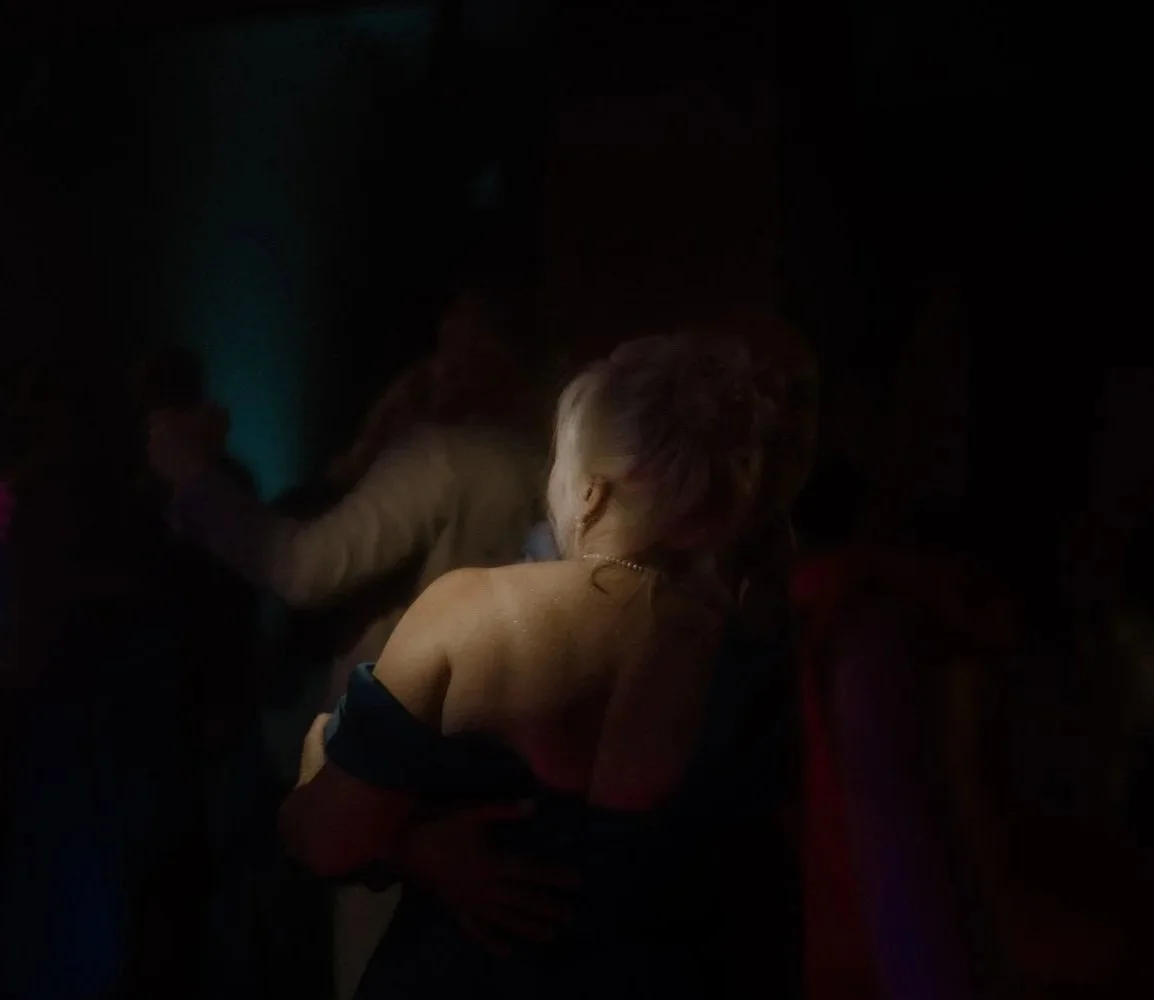 A woman in an off-the-shoulder dress dancing with a man in a dark environment, with other people around in a dimly lit setting.