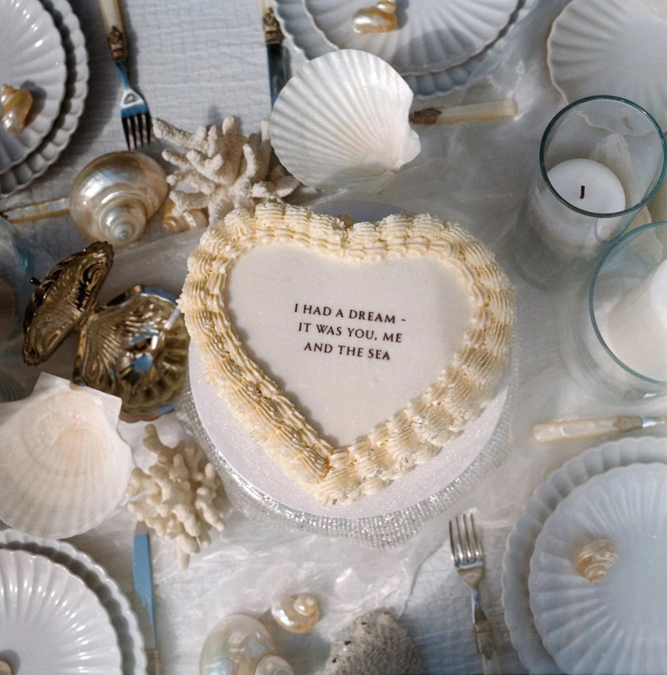 A heart-shaped white cake with a message that reads, 'I HAD A DREAM - IT WAS YOU, ME AND THE SEA,' surrounded by seashells, candles, and sea-themed decorations on a table.