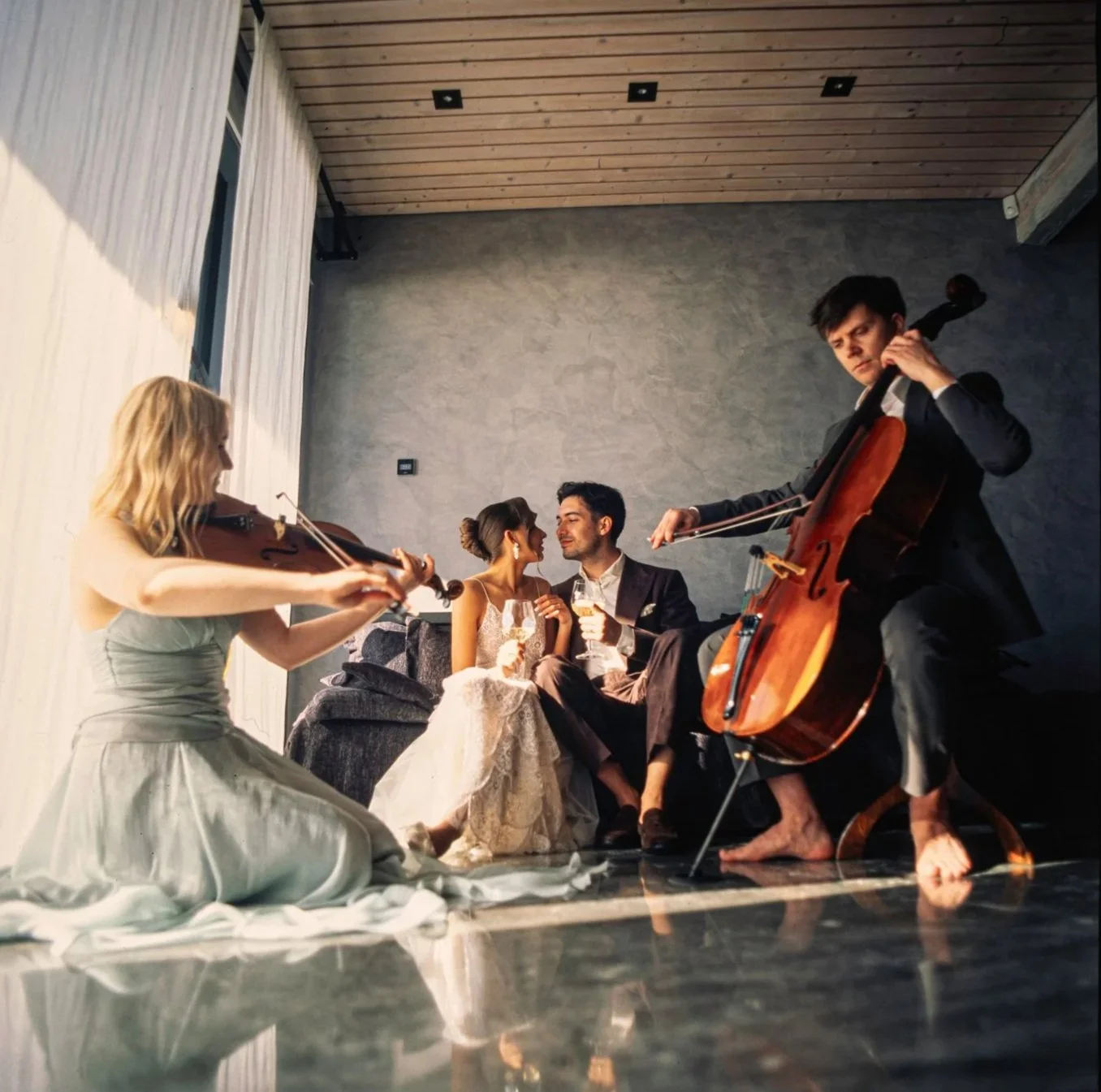 A couple is sitting on a sofa with two women playing musical instruments, a violin and a guitar, in a modern living room. The couple is smiling and holding glasses of wine, while the musicians are barefoot and focused on their instruments.