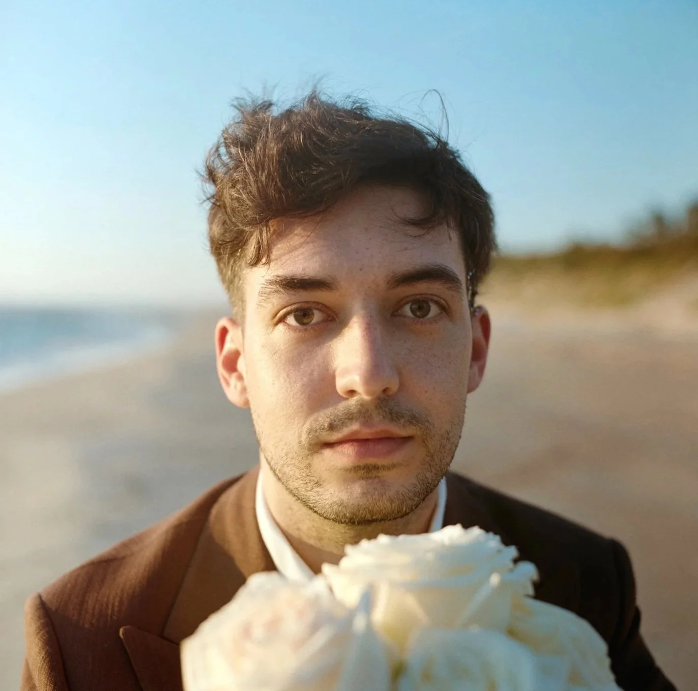 Young man with curly brown hair and light skin at the beach, holding a cupcake with white frosting, wearing a brown suit jacket