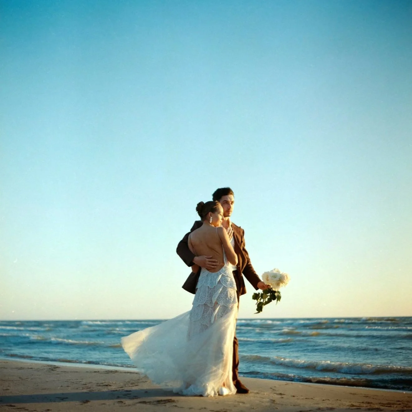 A couple in wedding attire standing on the beach at sunset, with the woman in a white wedding dress and the man in a brown suit, holding a bouquet of white roses.