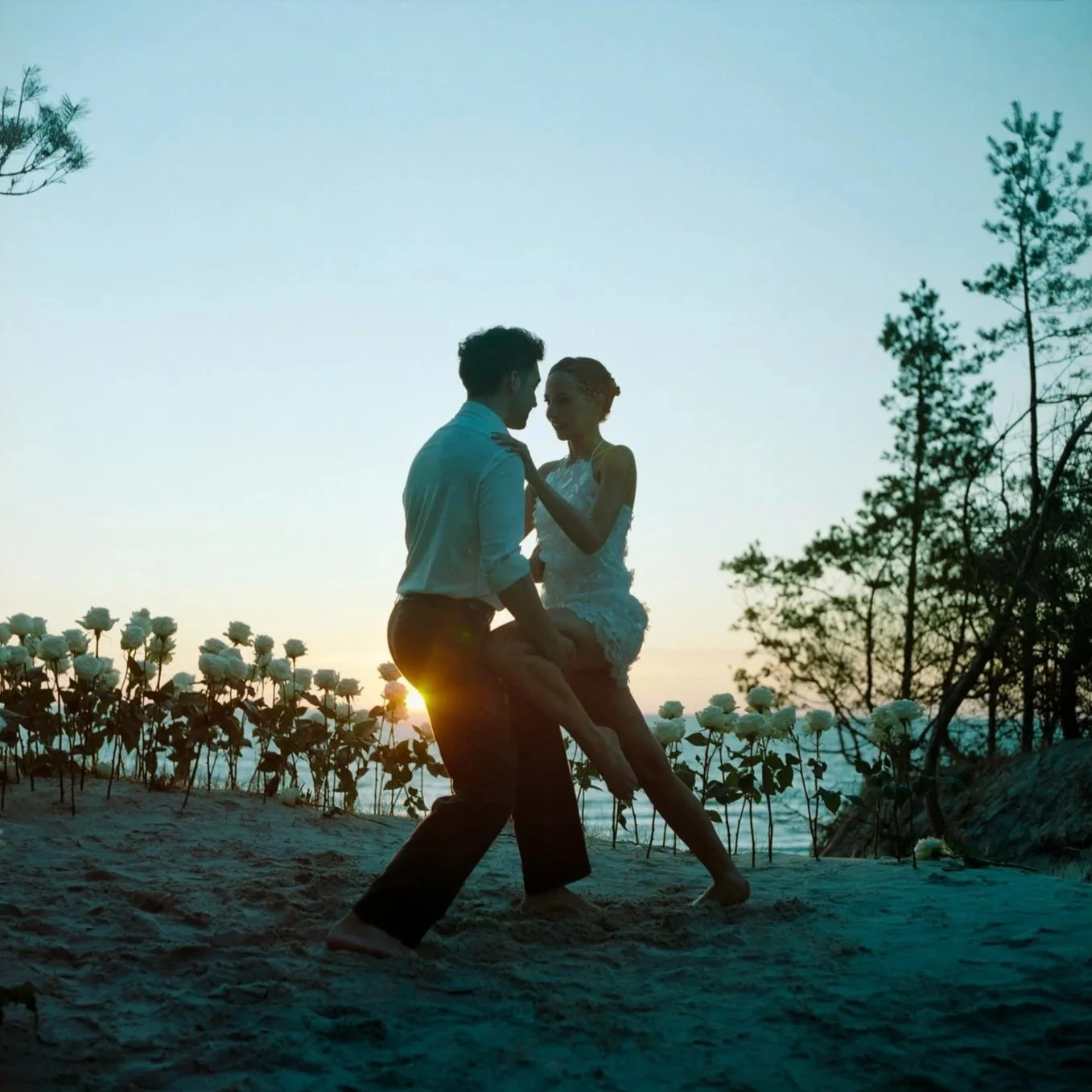 A couple dancing on the beach during sunset, surrounded by white roses and trees.