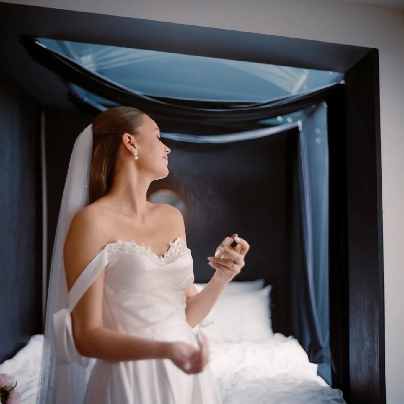 A bride in a white wedding dress with off-the-shoulder sleeves holding a ring box, sitting on a bed with dark curtains and pillow in a dimly lit room.