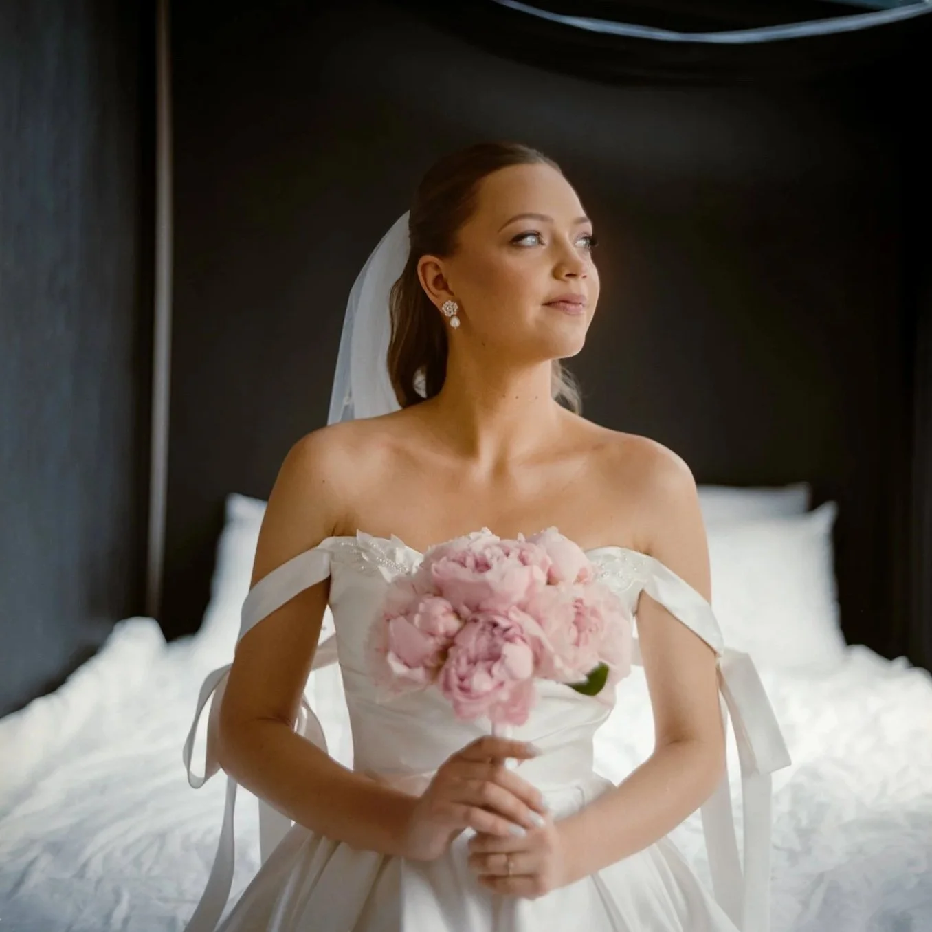 A bride wearing a white wedding dress with off-shoulder ribbons, holding a pink peony bouquet, sitting on a bed, looking thoughtfully to the side.