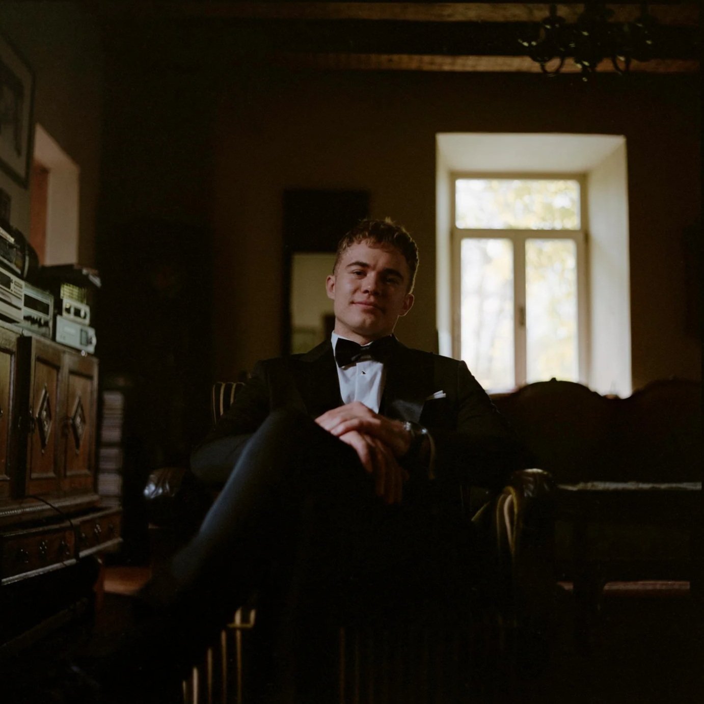 A young man dressed in a tuxedo, sitting confidently in a dimly lit room with a large window in the background.