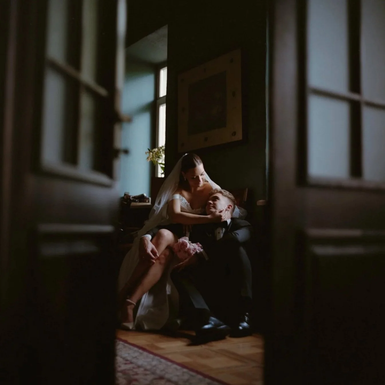 A bride sitting on a groom's lap in a dimly lit room, seen through an open door, with natural light coming from a window behind them.