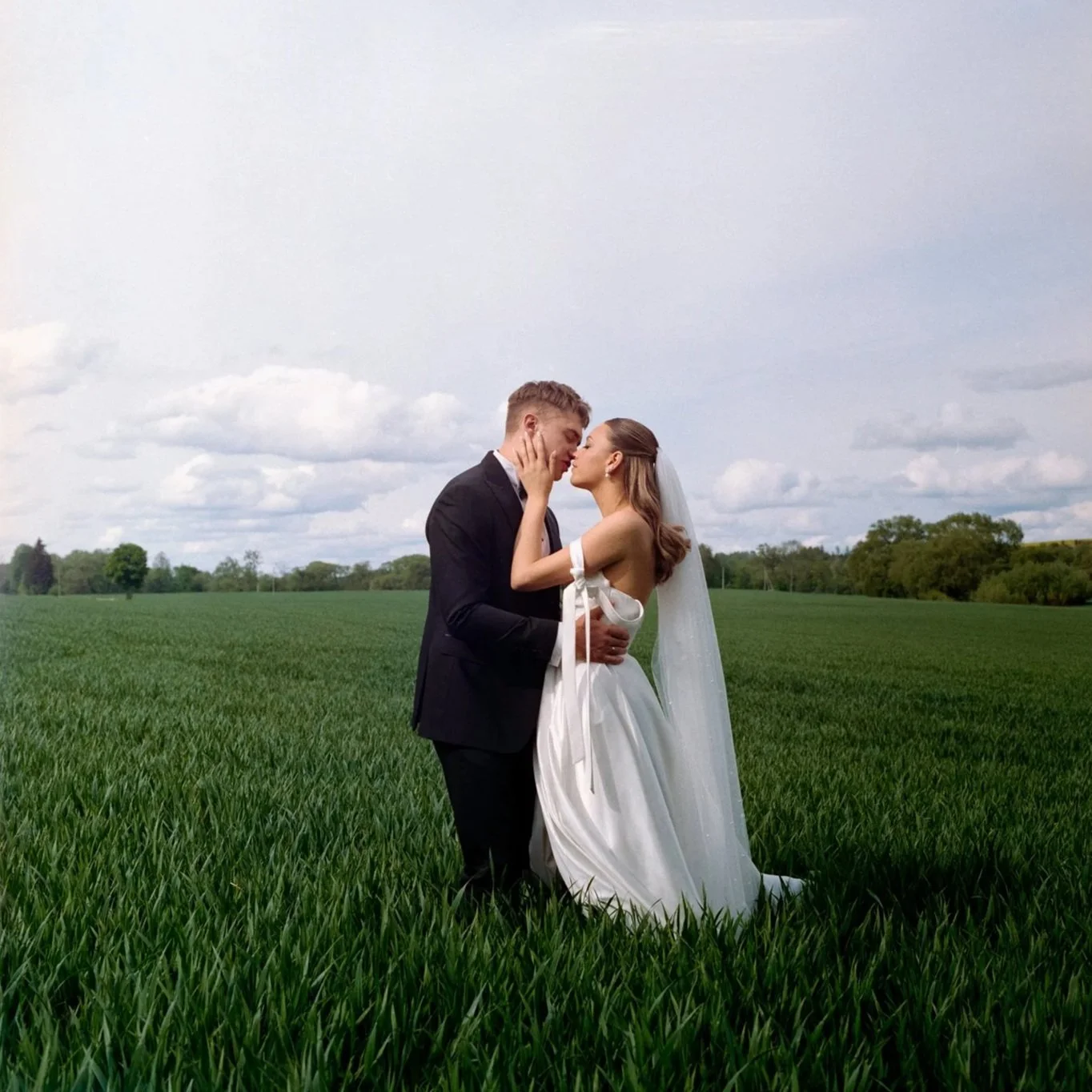 A bride and groom standing close together in a green field, about to kiss, with a cloudy sky overhead.