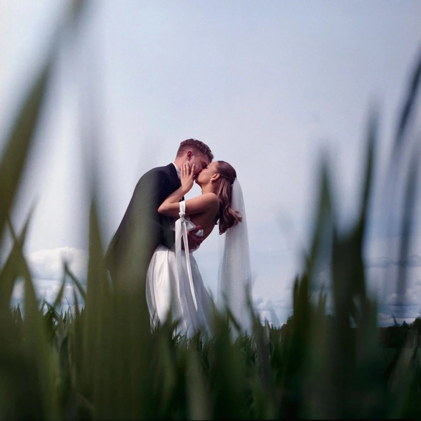 A newlywed couple kissing outdoors among tall grass, with sky and clouds in the background.