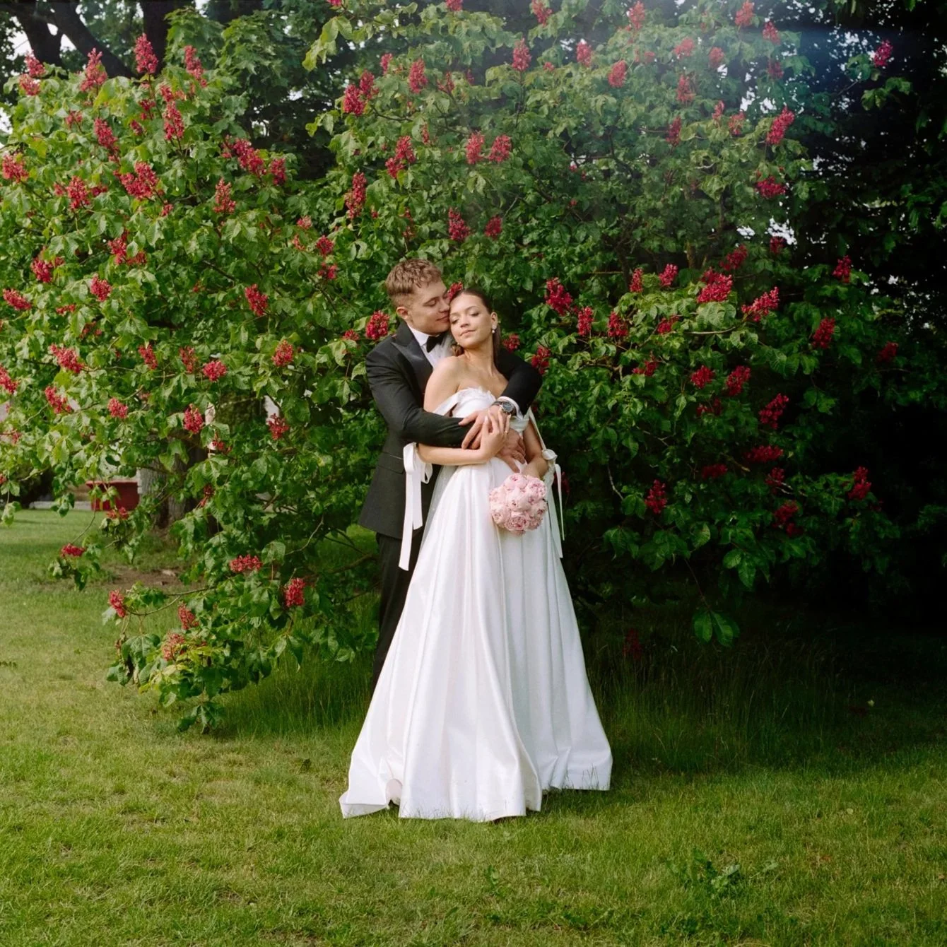 A bride and groom in wedding attire embracing outdoors in front of a leafy, flowering bush with red blossoms.