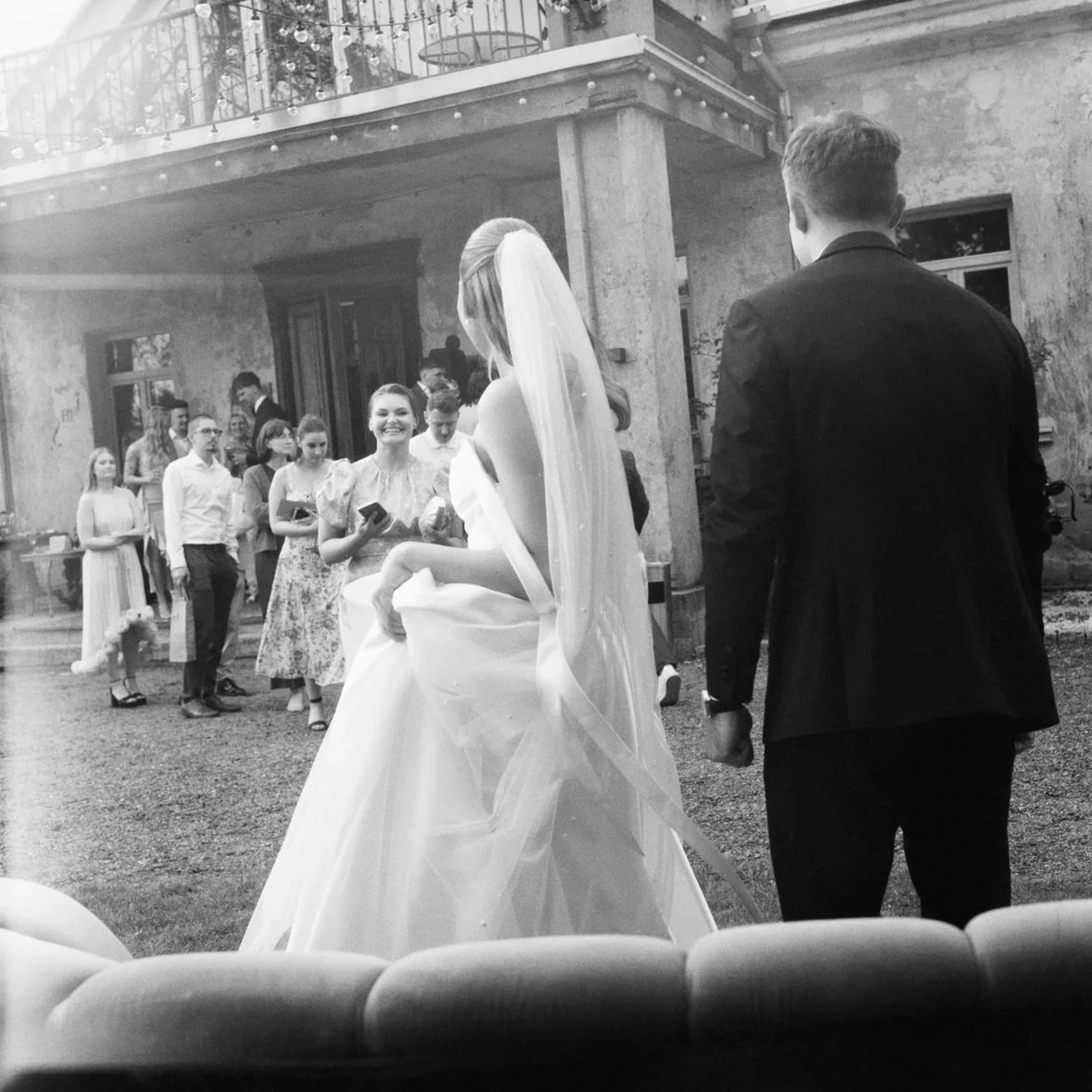 A black and white photo of a wedding scene showing a bride in a wedding dress and veil, and a groom in a suit, standing outdoors. Guests are in the background, some smiling and holding books or glasses.