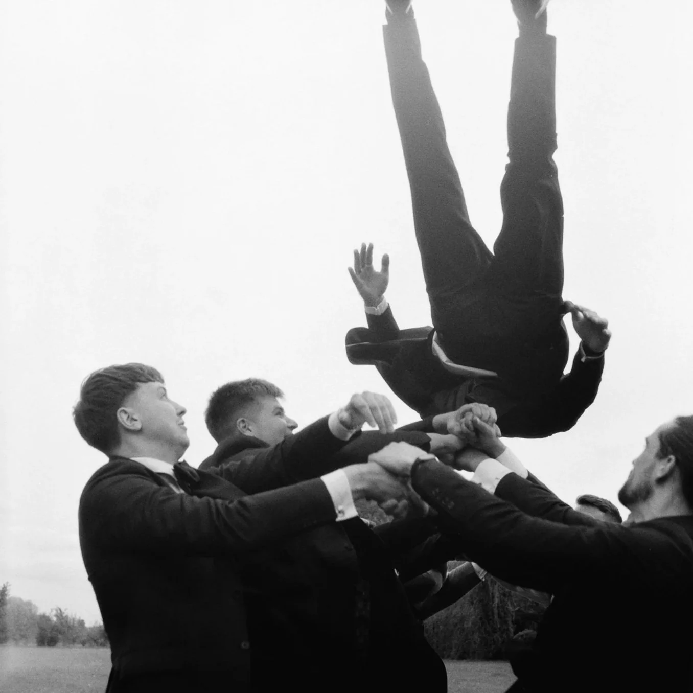 Group of men in suits participating in a human tower or pyramid formation outside, with one man upside down at the top, against a plain sky background.